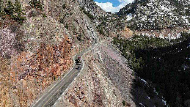 Semi Truck Driving on the edge of a cliff with a huge drop off, surrounded by mountains, pine trees at bottom of cliff, spring in Ouray Colorado, red mountains pass, Million Dollar Highway