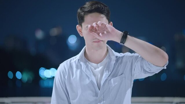 Portrait shot of an unhappy young man looking disappointed to camera with a city skyscraper view. A broken heart caucasian teenager guy try hard to stop crying in the cold night. Facial expression