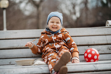 Cute toddler girl eating lunch in park, sitting on bench. Picnic in park during cold spring day.