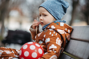 Cute toddler girl eating lunch in park, sitting on bench. Picnic in park during cold spring day.