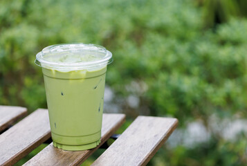 A green matcha tea drink in a plastic cup sits on a wooden table with garden background.