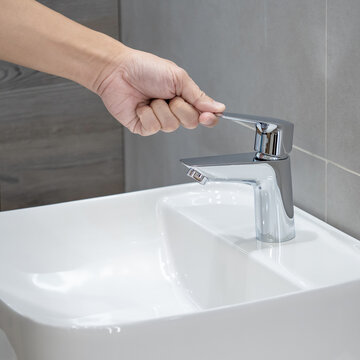 User hand holding a handle part of the chrome water tap over the ceramic wash basin at home interior showroom display. Choosing modern stainless steel water faucet for domestic bathroom design
