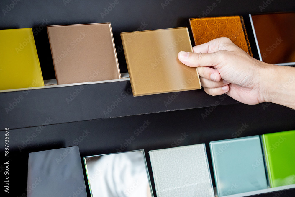 Wall mural architect hand choosing and picking at beige lacquered glass tile on material samples shelf display 