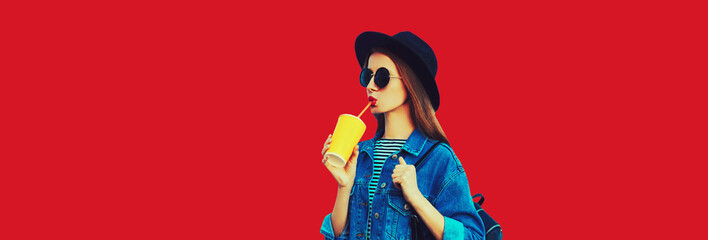 Stylish young woman drinking juice wearing black round hat, jean jacket on red background