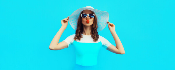 Portrait of beautiful young woman posing blowing kiss wearing summer straw hat on blue background