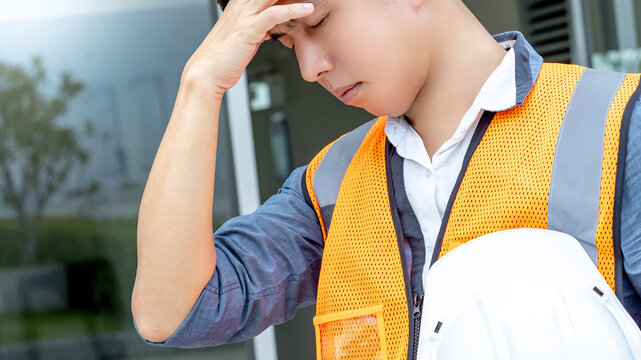 Dizziness and migraine headache from overworked exhaustion or heat stroke. Tired Asian male worker carrying safety helmet touching his forehead while working at construction site in extremely hot day