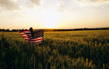 Young  woman with American flag in a wheat field at sunset celebrate Independence day. 4th of July....