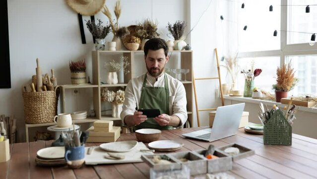 Bearded mature man potter takes photos of handmade bowl from clay for website of his own pottery shop. Caucasian male updates web page and social media portfolio.