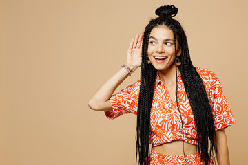 Young curious nosy smiling Latin woman she wears orange casual clothes try to hear you overhear listening intently isolated on plain pastel light beige background studio portrait. Lifestyle concept.