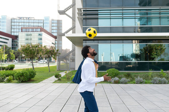 Happy businessman holding blazer and playing with soccer ball in office park