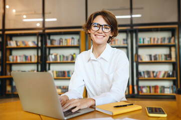 Young confident happy successful employee IT business woman wear white shirt casual clothes glasses hold use work on laptop pc computer surfing internet sit at office desk. Achievement career concept.