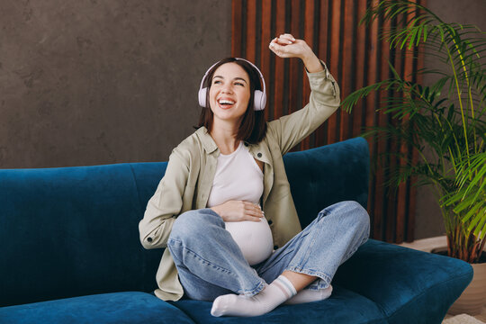Young pregnant woman with belly wear casual clothes listen to music in headphones sit on blue sofa couch stay at home hotel flat rest relax spend free time in living room. Maternity pregnancy concept.