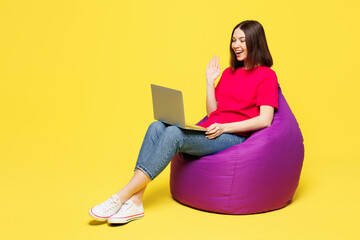 Full body young IT woman wear pink t-shirt casual clothes sit in bag chair hold use work on laptop pc computer waving hand isolated on plain yellow orange background studio portrait Lifestyle concept