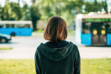 Rear view of adult brunette female standing on the street and looking at traffic and buses passing...