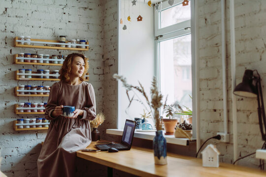Businesswoman sitting with coffee cup near laptop on desk in workshop
