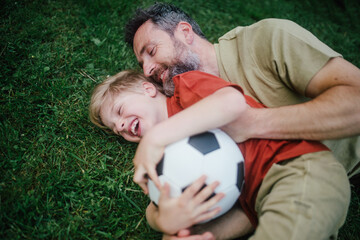 Dad lying on grass with son, laughing and having fun. Father and boy playing football, catching...
