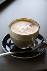 One transparent cup of cappuccino coffee on black plate with spoon and dark background. Artistic picture. Single object. Cappuccino design with small heart. Tranquility morning pattern. Copyspace