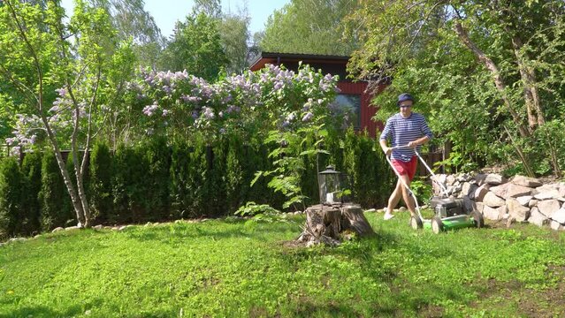 Middle-aged man trimming overgrown green lawn with lawn mower in garden. Male gardener in shorts using lawn trimmer cutting grass while landscaping. Concept of seasonal work.