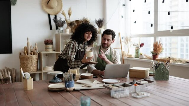 Caucasian couple of co-workers work on small pottery production business. Mature man and young curly-haired woman in aprons use laptop to take inventory of products on store website.