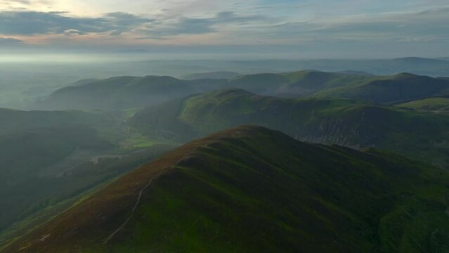 Ancient rounded glaciated forested mountains and fells misting into lowlands at golden hour. From Grisedale Pike, Lake District, Cumbria, England, UK