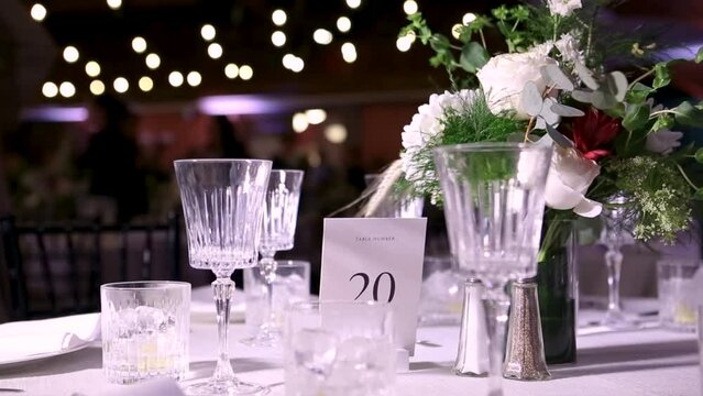 Close-up of an elegant table setting featuring crystal glasses, a floral centerpiece, and a table number card at a wedding reception with soft, romantic lighting. Wedding Reception, USA