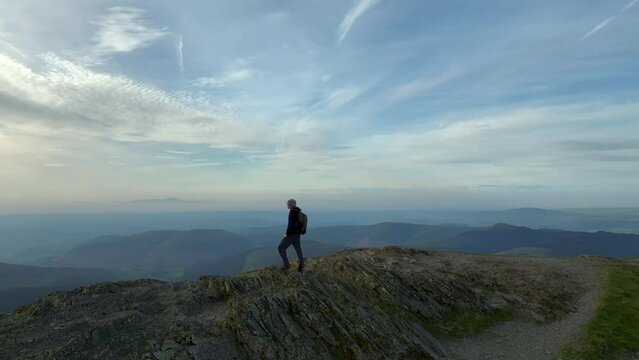 Mountain walker on craggy summit at golden hour. Camera 180 orbit revealing misty mountains and wide green valley with lake. Grisedale Pike, Lake District, Cumbria, England, UK