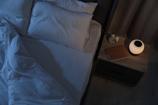 Nightlight, alarm clock and book on bedside table near bed indoors, above view