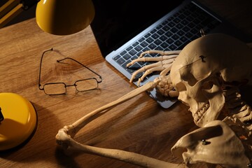 Waiting concept. Human skeleton sleeping at wooden table with laptop