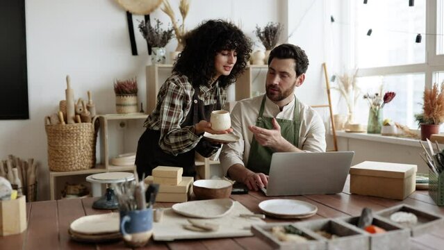 Caucasian couple of co-workers work on small pottery production business. Mature man and young curly-haired woman in aprons use laptop to take inventory of products on store website.