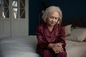 Unhappy sad elderly woman wearing pajamas sitting on bed feeling loneliness