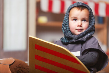 Toddler dressed up as a knight playing with shield in room game. Home game, medieval.