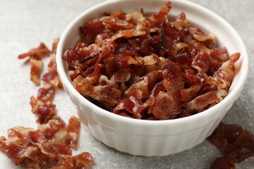 Pieces of tasty fried bacon in bowl on gray textured table, closeup