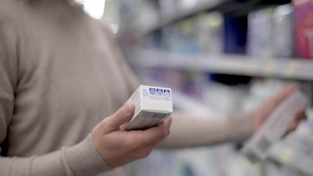 A woman holds two toothpaste boxes, comparing their details in a store aisle. The setting is a well-lit retail environment with various dental care products on shelves.
