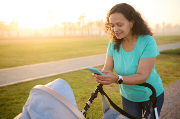 Obraz premium Happy mom shopping online, taking photo of her newborn child sleeping in baby buggy. Young pretty woman, mother using mobile phone while pushing baby pram with her baby, standing outdoors at sunset