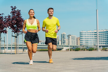 Two joggers in bright athletic wear running on a sunny day with a modern cityscape in the background.