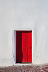 A vibrant red door stands alone in the center of a white wall, casting a slight shadow. The design is simple and clean, highlighting the door as the focal point.