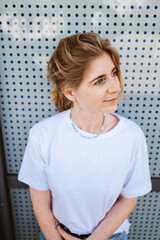 Portrait of a pensive woman standing against a perforated wall, looking off into the distance. She...