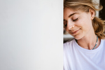 Serene young woman leaning against a white wall with eyes closed, exuding relaxation and peace.
