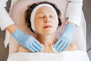 Mature woman lying in cosmetic clinic, doctor in latex gloves performing cosmetic procedures