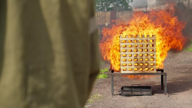 Fire training exercise in which firefighter practices extinguishing fire in simulated structure. Flames are intense. Firefighter is seen. Fire burns brightly and flames lick up sides of structure.