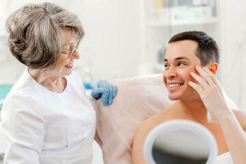 Happy man and senior smiling female cosmetologist doctor talking, looking in mirror