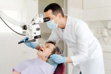 Young dentist doctor, man treating teeth using dental microscope, checking senior attractive woman