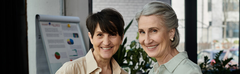 Mature, beautiful lesbian couple standing side by side in an office setting.