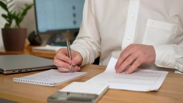 Unrecognizable man flips stack of papers with financial indicators, businessman writes with pen in notebook, making economic notes close up. Male executive using laptop for remote business studying