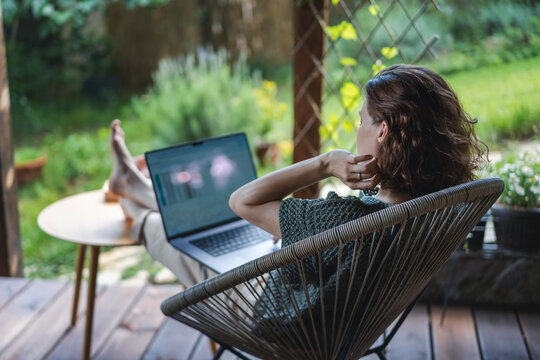 Young adult Caucasian woman sitting on cottage terrace using laptop in summertime, online work