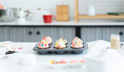 Cupcakes with pastel color butter cream, colorful jelly and chocolate flakes on baking tray in kitchen at home. Flour, sugar and decorating candy on table. Selective focus