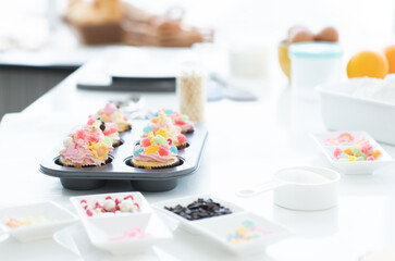 Cupcakes with pastel color butter cream, colorful jelly and chocolate flakes on baking tray in kitchen at home. Flour, sugar, eggs and decorating icing candy on table. Selective focus