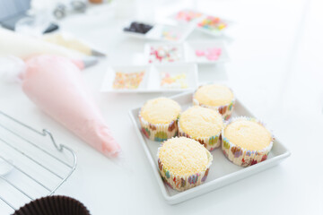 Selective focus on fresh cakes baked from oven. Piping bags with pastel butter cream on table with ingredients and decorations of homemade cupcakes on table in kitchen at home