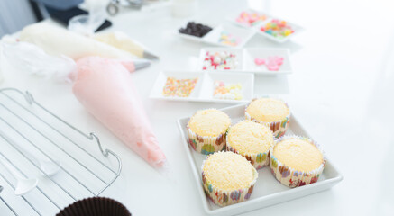 Selective focus on fresh cakes baked from oven. Piping bags with pastel butter cream on table with ingredients and decorations of homemade cupcakes on table in kitchen at home