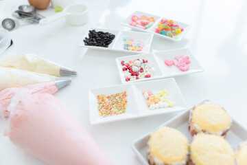 Selective focus on icing candy, chocolate flakes or coated sugar and jelly in tray for decorating. Fresh cupcakes baked from oven, piping bags with pastel butter cream, ingredients on white table